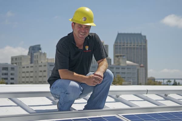 Photo of Daniel Sullivan wearing hard hat and kneeling next to rooftop solar array with downtown San Diego in background