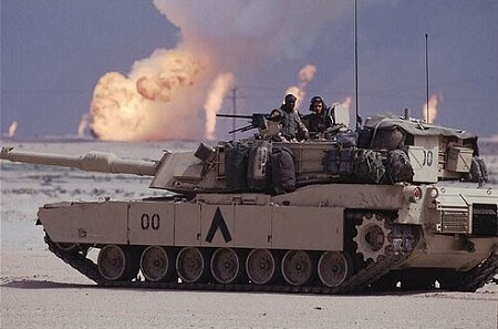 Image of troops riding a tank through dessert with burning oil field in background