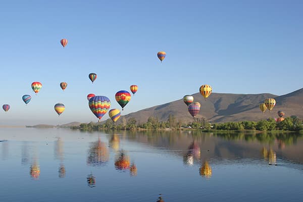 Photo of brightly colored hot air balloons at low altitude over lake with hills in background